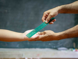 Illustrating corporal punishment, a teacher swats a student's hand with a plastic ruler.